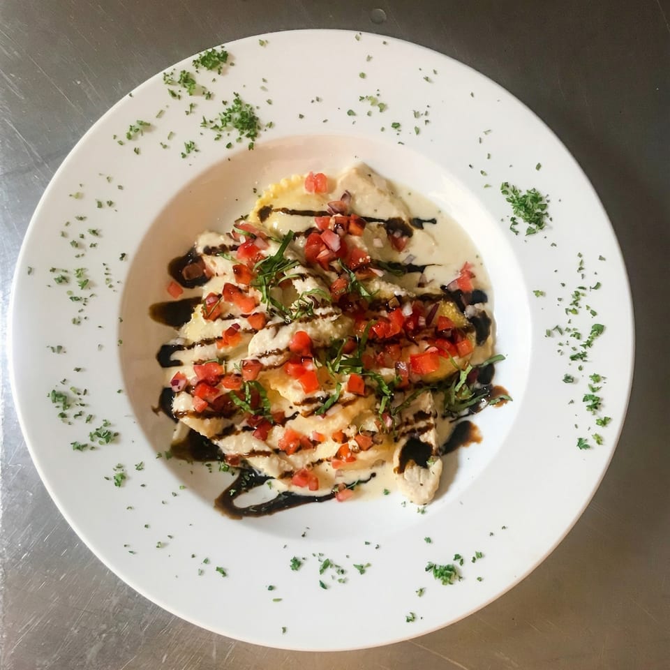 Ravioli in cream sauce with diced tomatoes, basil, and balsamic glaze on a white plate.